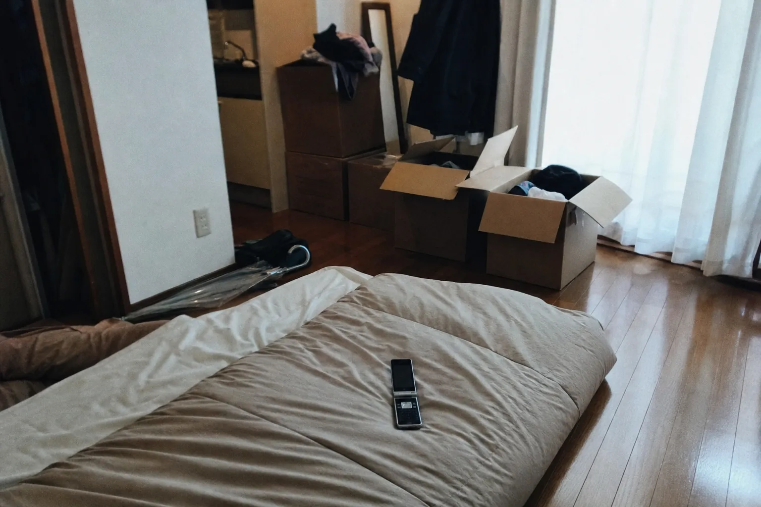 昼下がりの狭い部屋、畳んだ布団の上に開いたままのガラケー