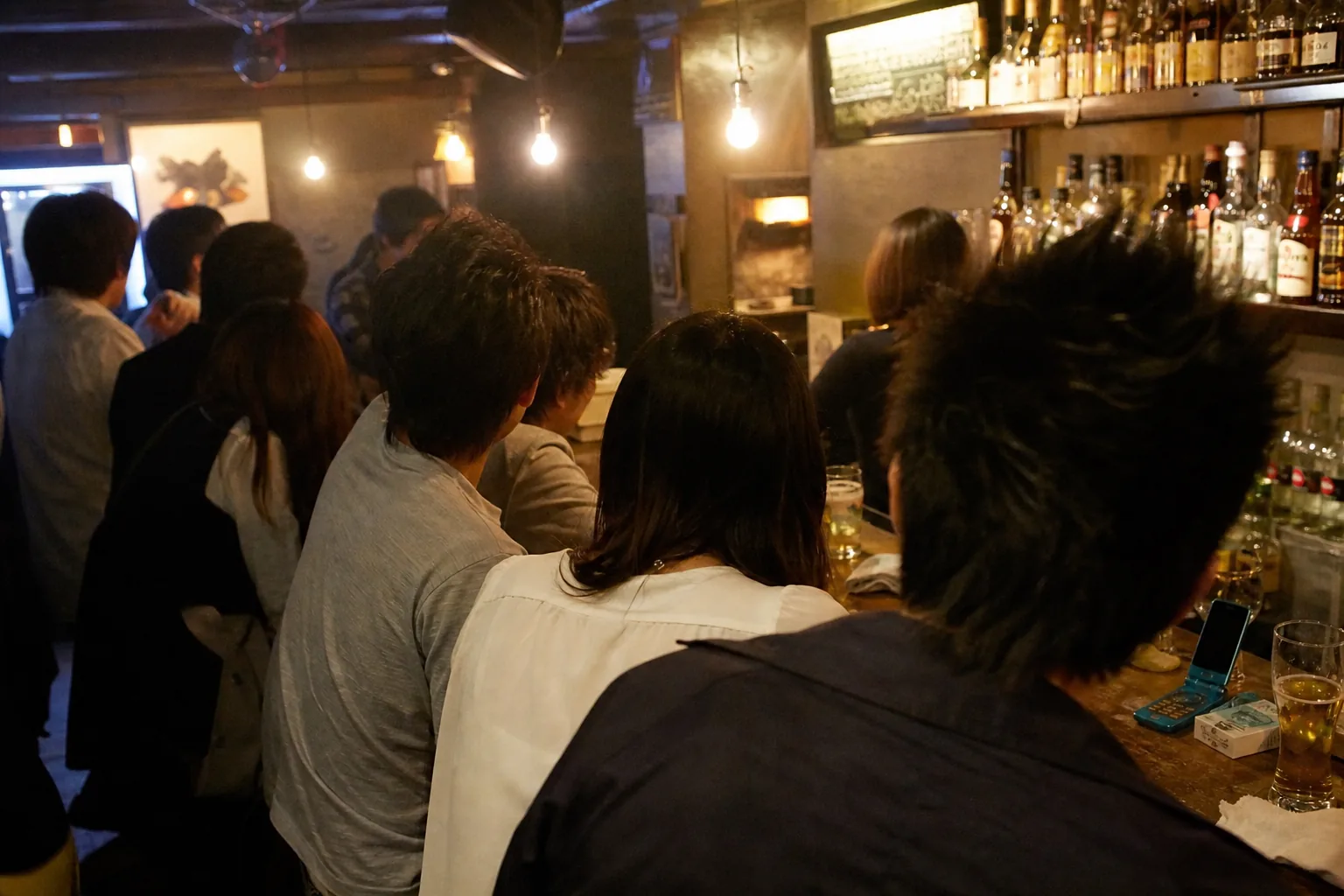 夜の都心のバー、オフ会の10人ほどを最後列の背中側から。全員の後ろ頭と肩だけ