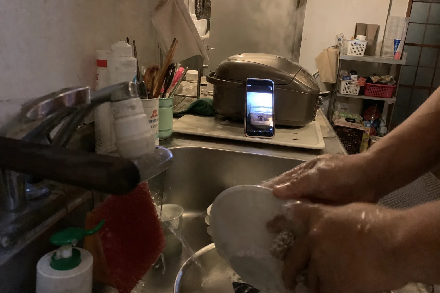 炊飯器に立てかけたスマホが光り、手前でシンクの皿を洗う手元、夕方
